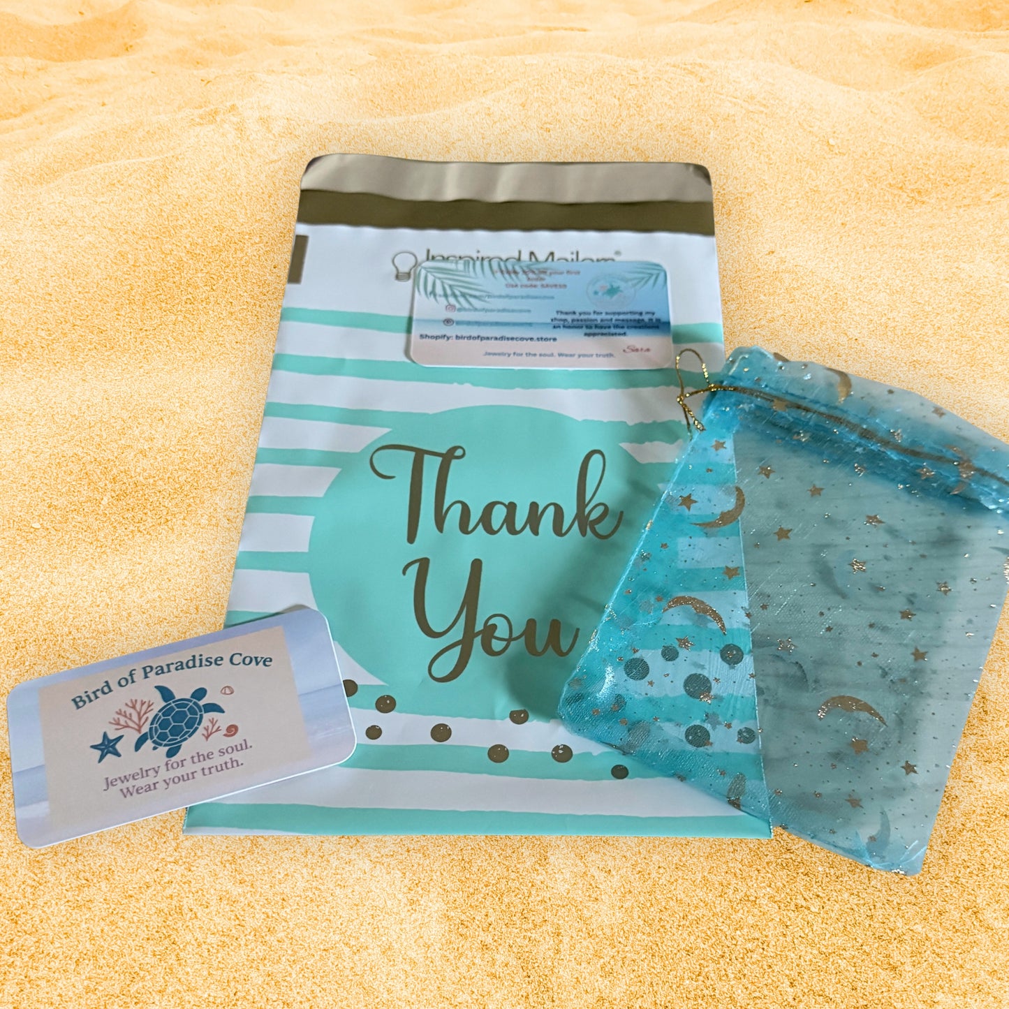 Bird of Paradise Cove handmade bracelet packaging with teal thank you mailer, celestial blue gift bag, and branded jewelry card on beach sand background