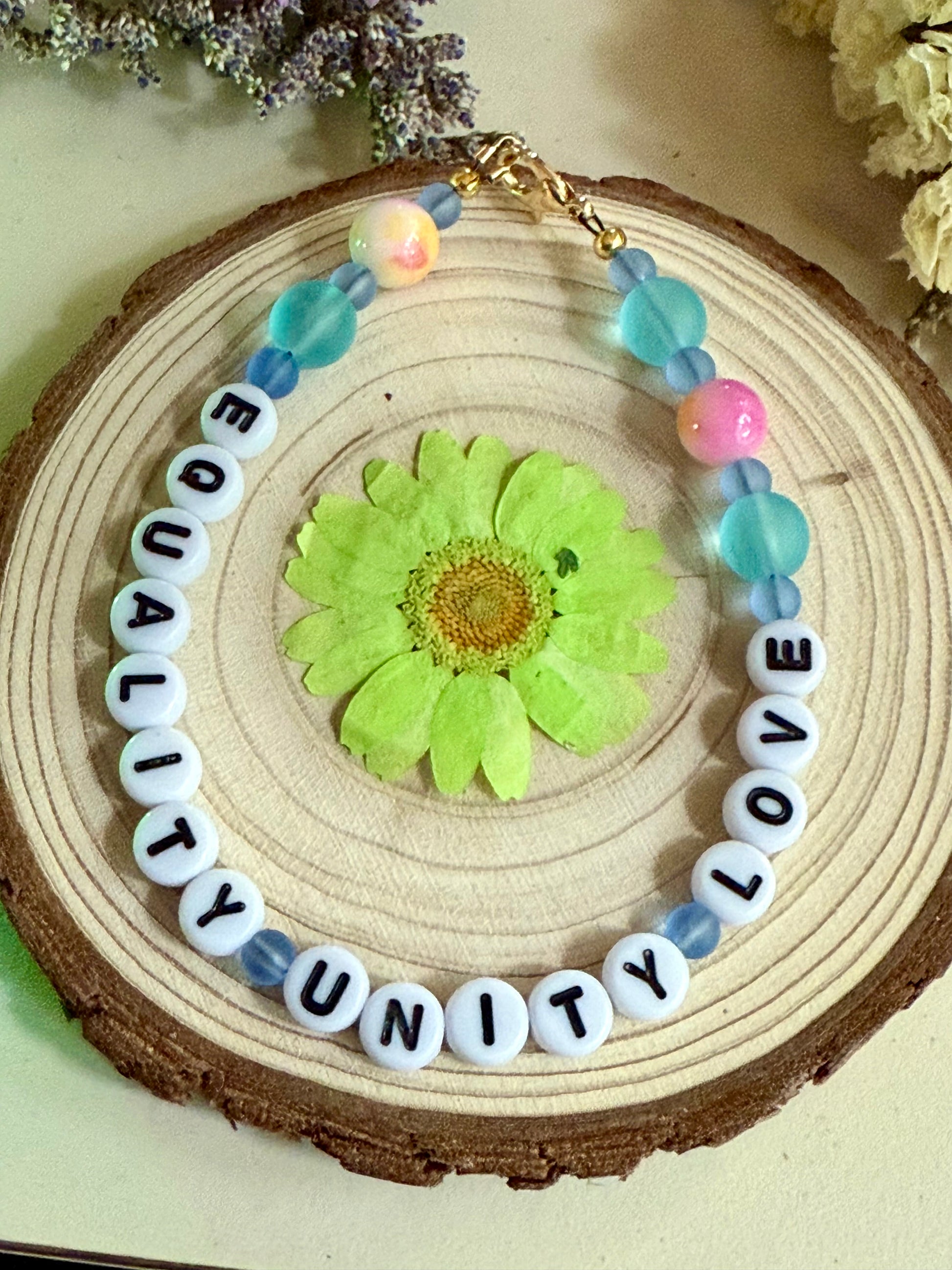 Handmade beaded bracelet spelling “Equality Love Unity” displayed on a natural wood slice with a pressed green flower and soft floral accents.