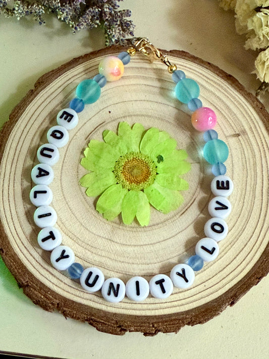 Handmade beaded bracelet spelling “Equality Love Unity” displayed on a natural wood slice with a pressed green flower and soft floral accents.
