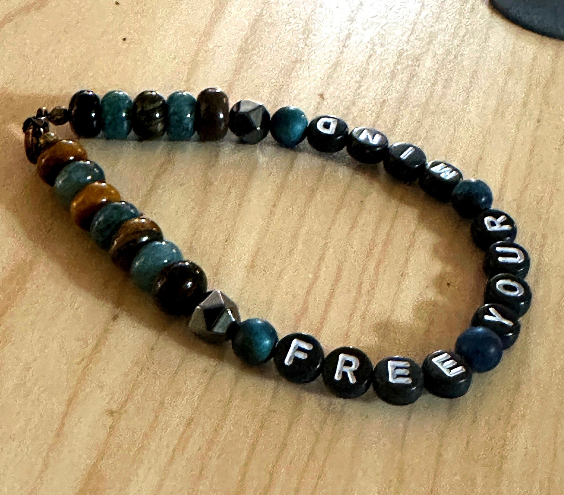 Handmade Free Your Mind beaded bracelet on natural wood slice with teal, brown, and black gemstones