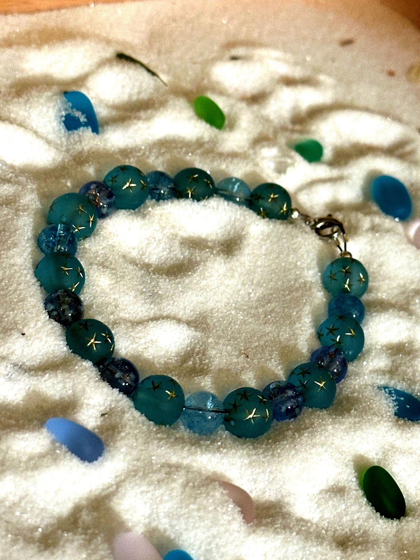 Coastal-inspired flat lay of a blue and teal beaded bracelet styled with seashells and sea glass on sand