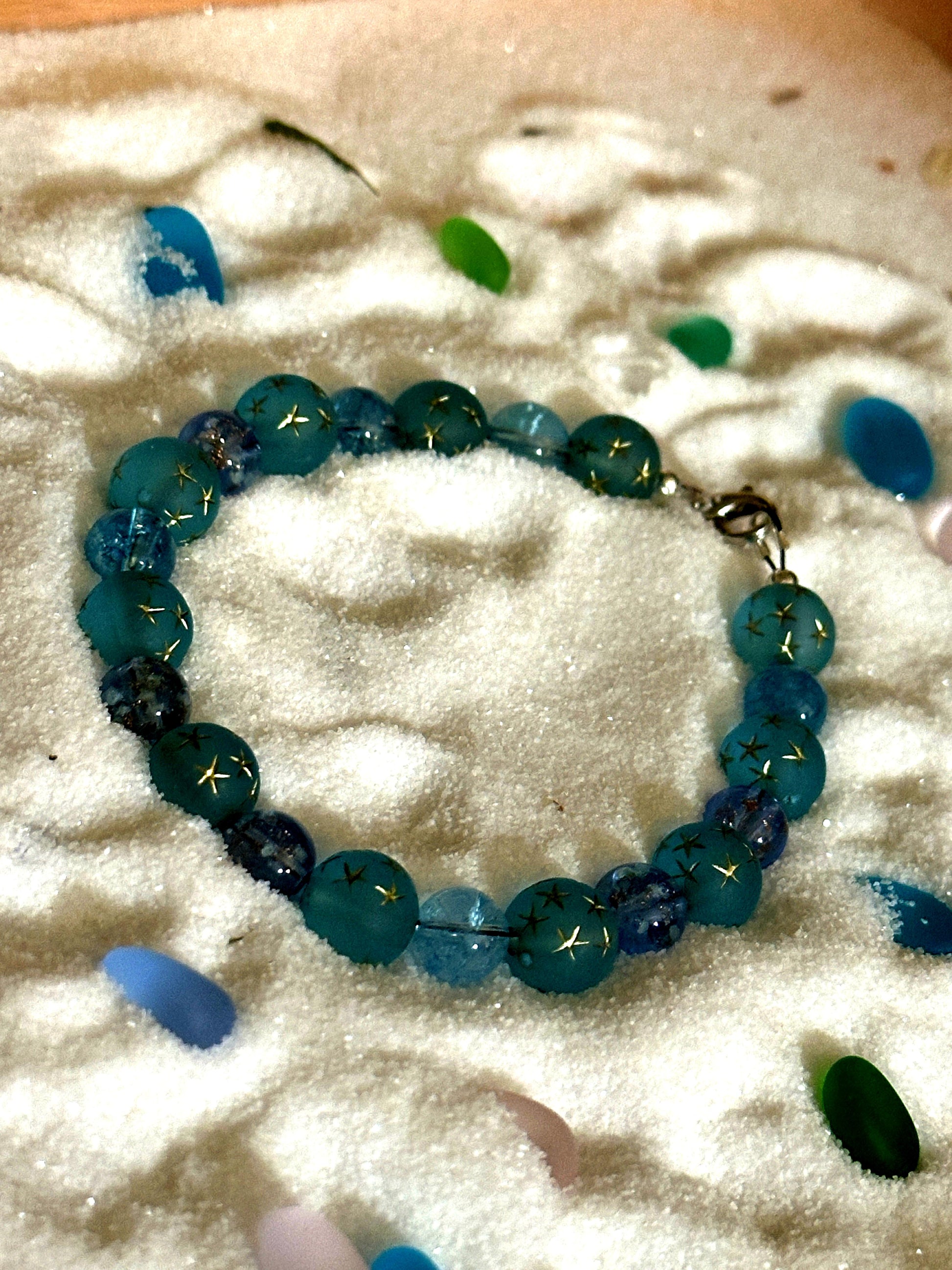 Coastal-inspired flat lay of a blue and teal beaded bracelet styled with seashells and sea glass on sand