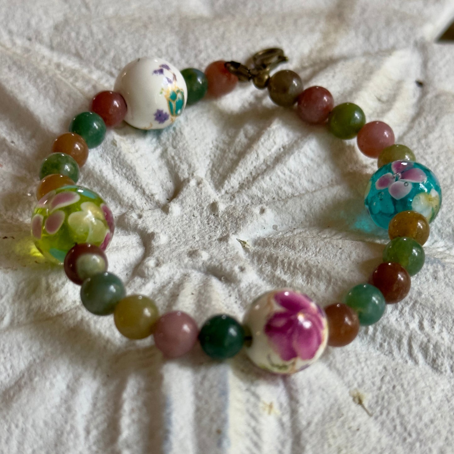 Handmade floral gemstone bracelet with multicolored beads and glass flower accents arranged in a circular layout on a textured white background.