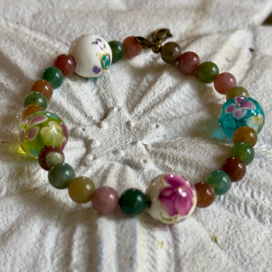 Handmade floral gemstone bracelet with multicolored beads and glass flower accents arranged in a circular layout on a textured white background.
