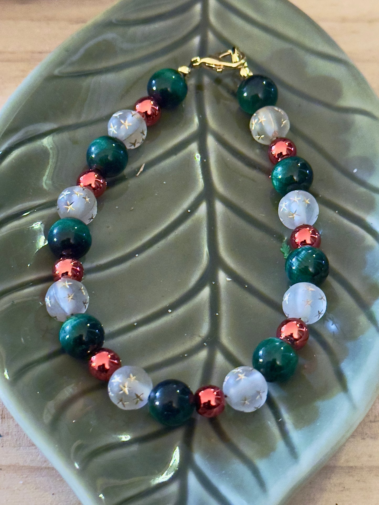 Handmade Christmas gemstone bracelet displayed on a green leaf dish, featuring deep green beads, red accents, and star-etched white beads with gold clasp.