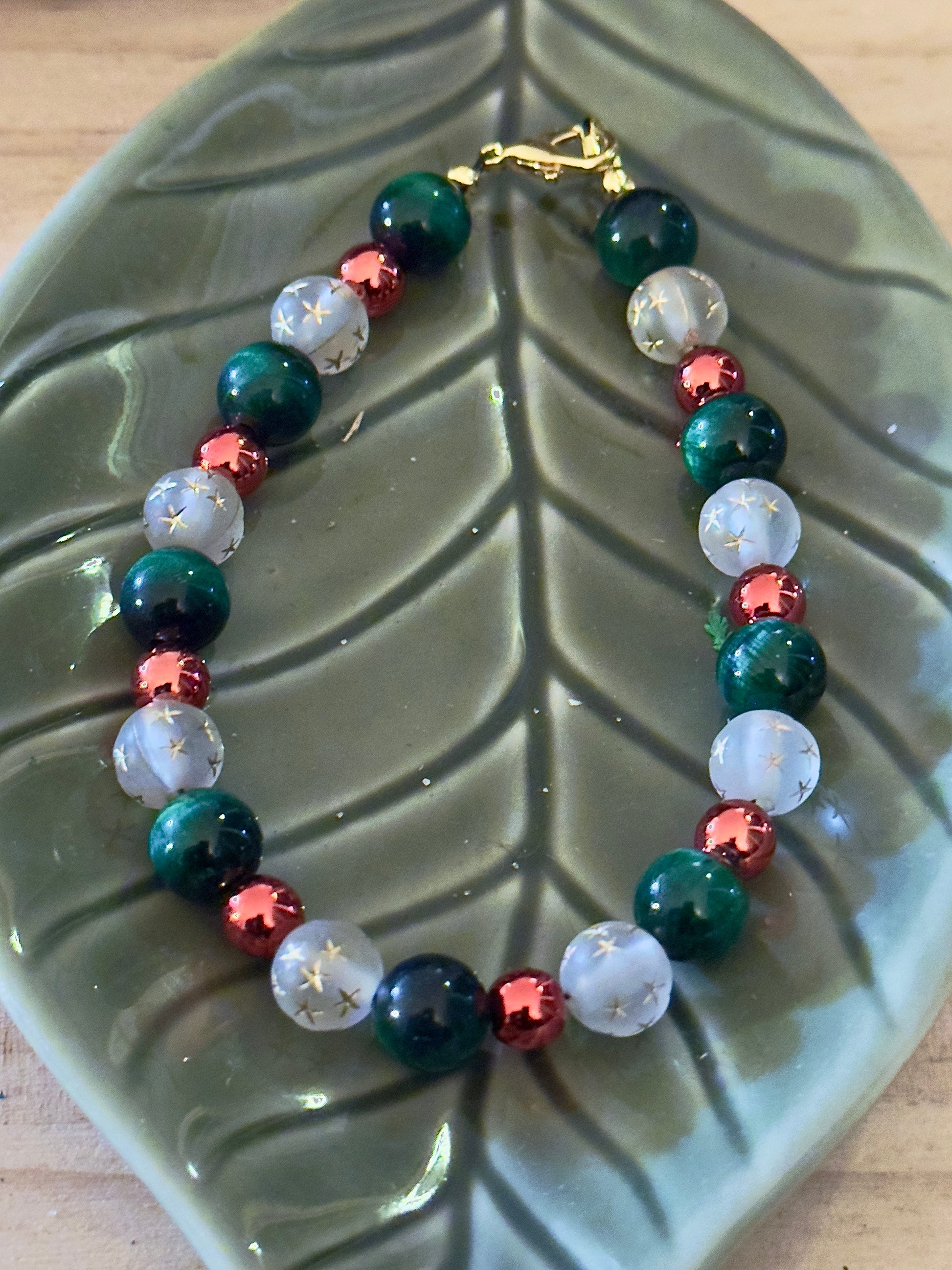Handmade Christmas gemstone bracelet displayed on a green leaf dish, featuring deep green beads, red accents, and star-etched white beads with gold clasp.