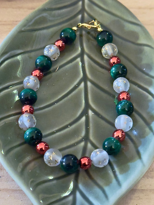 Handmade Christmas gemstone bracelet displayed on a green leaf dish, featuring deep green beads, red accents, and star-etched white beads with gold clasp.