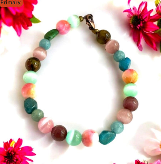 Colorful handmade gemstone bracelet arranged in a circle on a white background with bright pink and yellow flowers, featuring pastel pink, aqua, blue, and earth-tone beads with a bronze clasp.