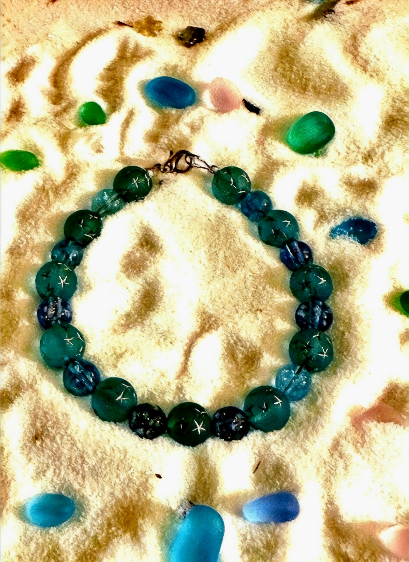 Handmade blue and teal beaded bracelet arranged in a circle on sand with scattered sea glass, coastal flat lay jewelry photo