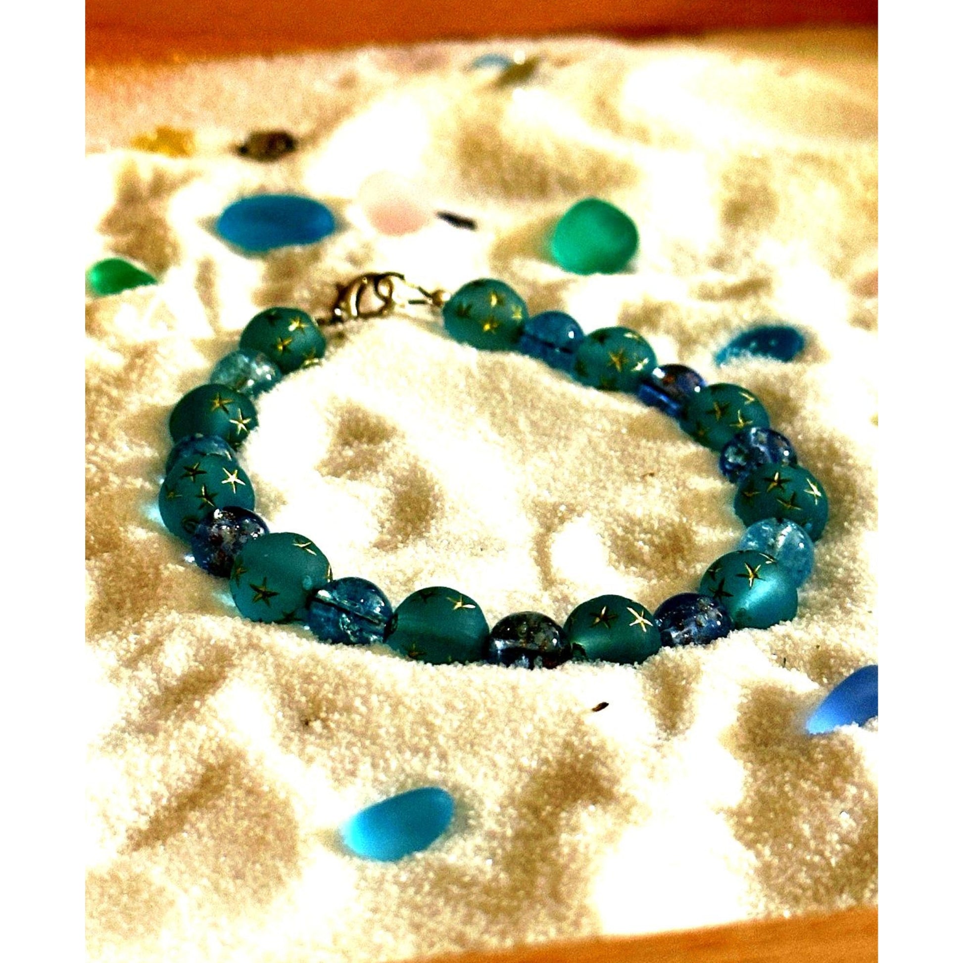 Handmade beaded bracelet in shades of blue and teal resting on sand with sea glass accents, coastal jewelry styling