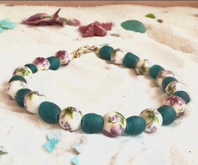 Nature-inspired beaded bracelet with floral ceramic and teal glass beads resting in sand with sea glass accents, calming handmade jewelry