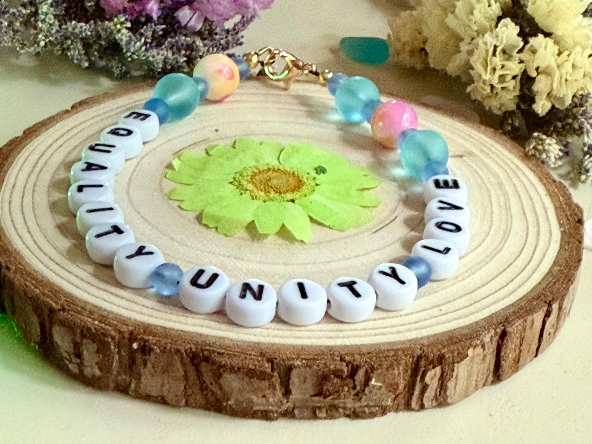 Equality Love Unity beaded bracelet on a wooden slice with green floral detail, styled in a gentle, earthy flat lay.