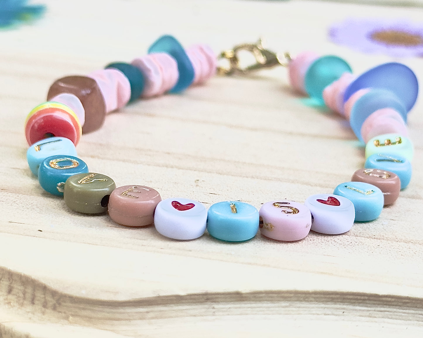 Lifestyle flat lay of a handmade “LOVE IS LOVE” bracelet styled with soft florals, fabric, and calming pastel decor.