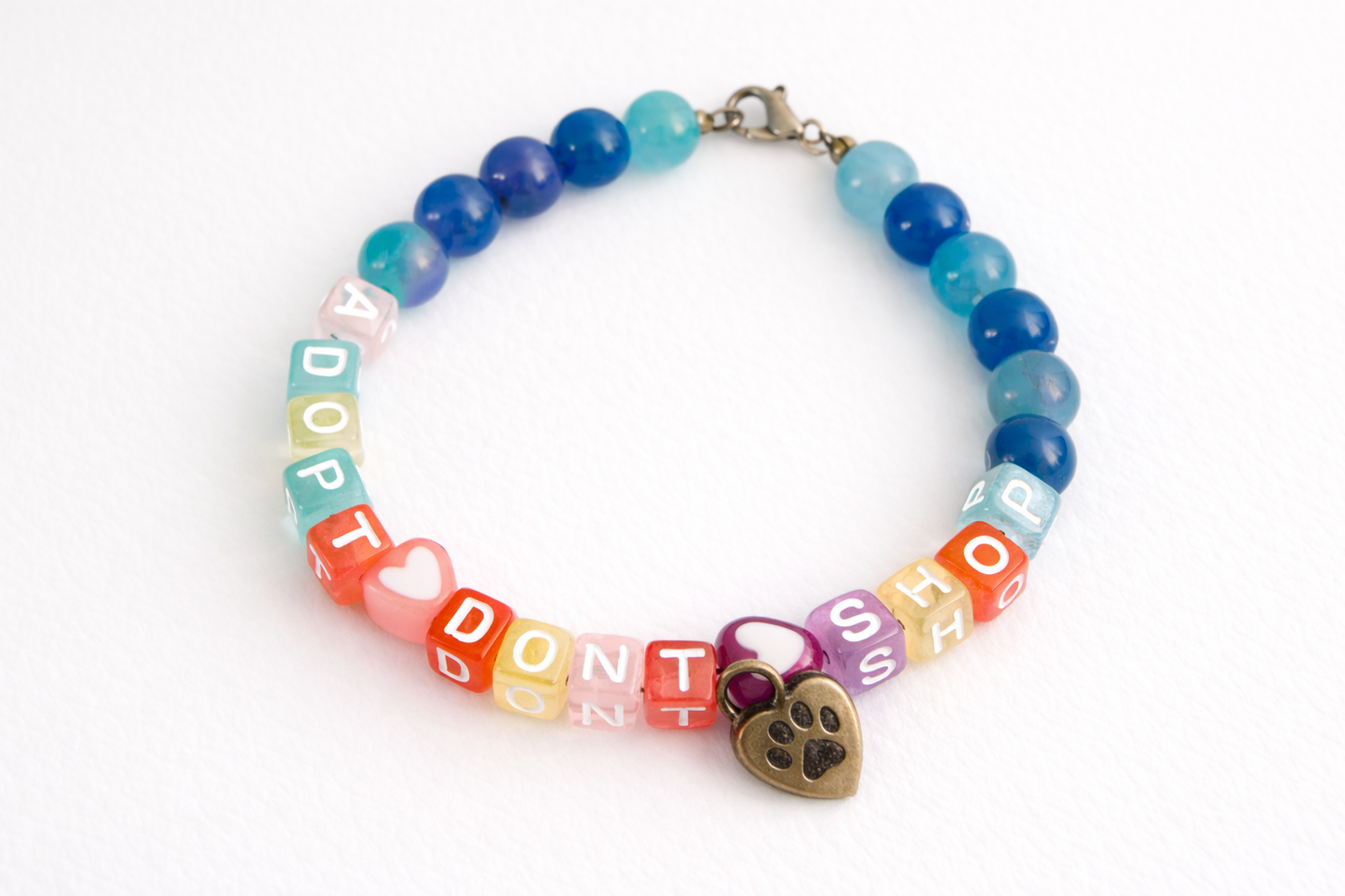 Handmade “Adopt Don’t Shop” beaded bracelet with blue and teal gemstone beads, colorful letter beads, heart accents, and paw print charm on a clean white background.
