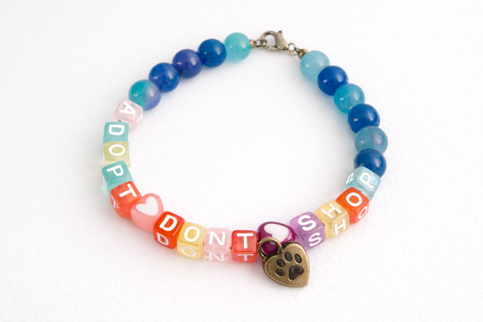 Handmade “Adopt Don’t Shop” beaded bracelet with blue and teal gemstone beads, colorful letter beads, heart accents, and paw print charm on a clean white background.
