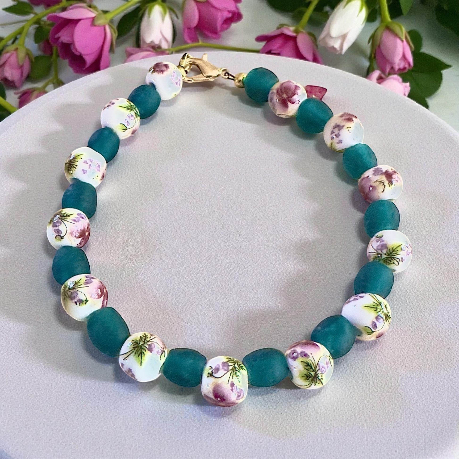 Floral ceramic and teal glass beaded bracelet with gold clasp, displayed on a neutral plate with pink flowers, handmade nature-inspired jewelry