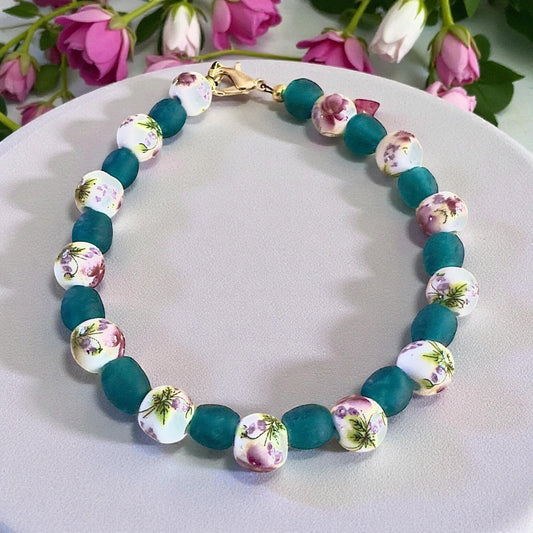 Floral ceramic and teal glass beaded bracelet with gold clasp, displayed on a neutral plate with pink flowers, handmade nature-inspired jewelry