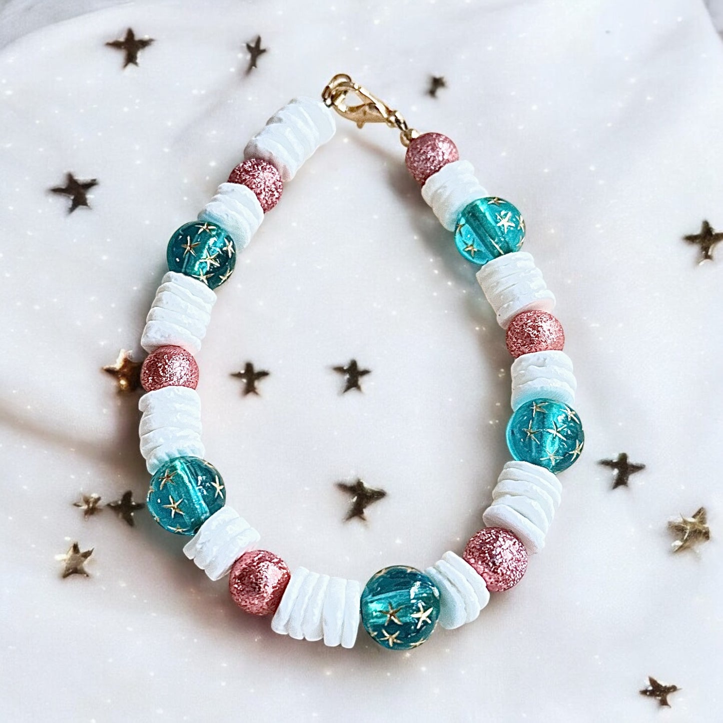 Ocean-inspired beaded bracelet with teal star beads, white shell spacers, and rose shimmer accents photographed on a light background with subtle star details.