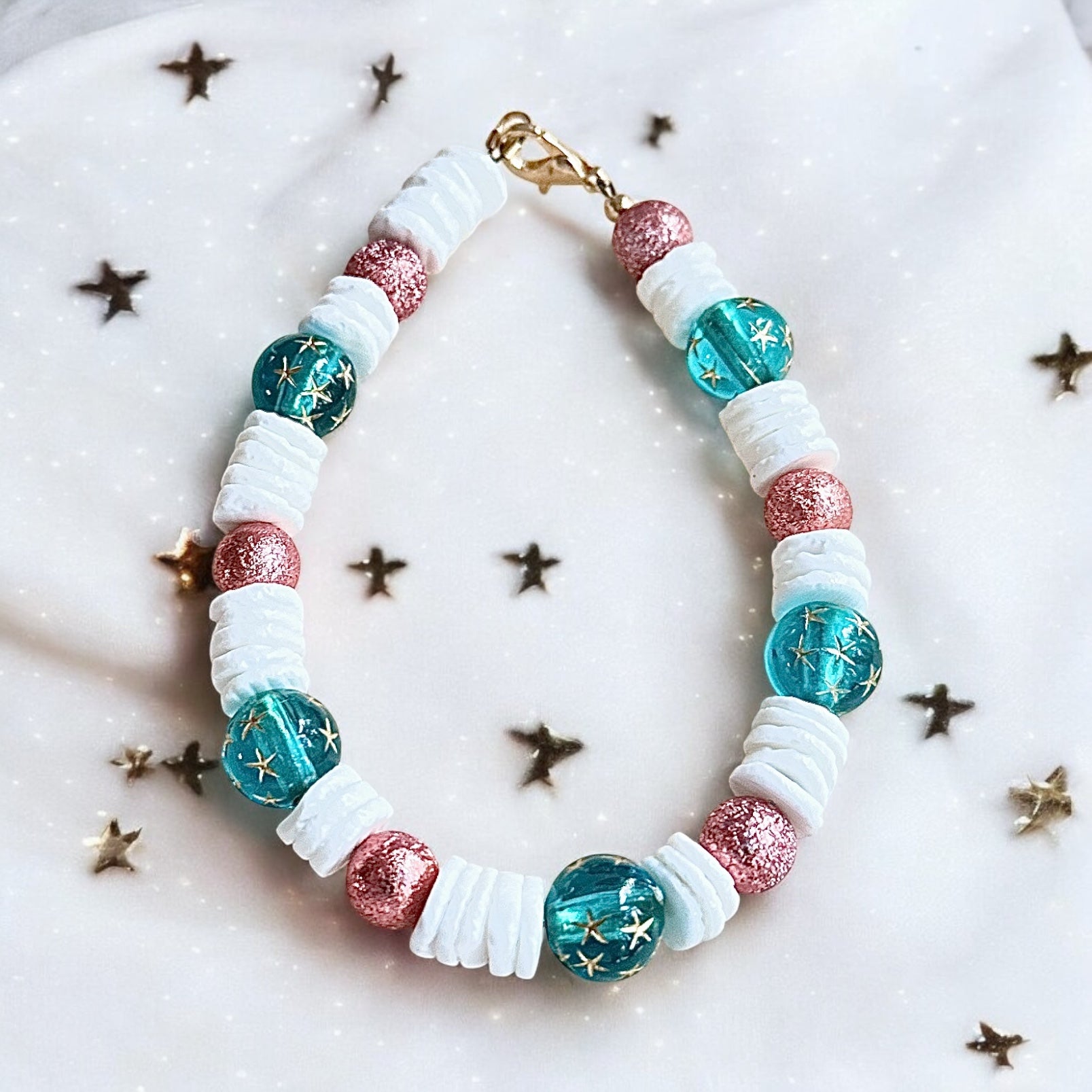 Ocean-inspired beaded bracelet with teal star beads, white shell spacers, and rose shimmer accents photographed on a light background with subtle star details.