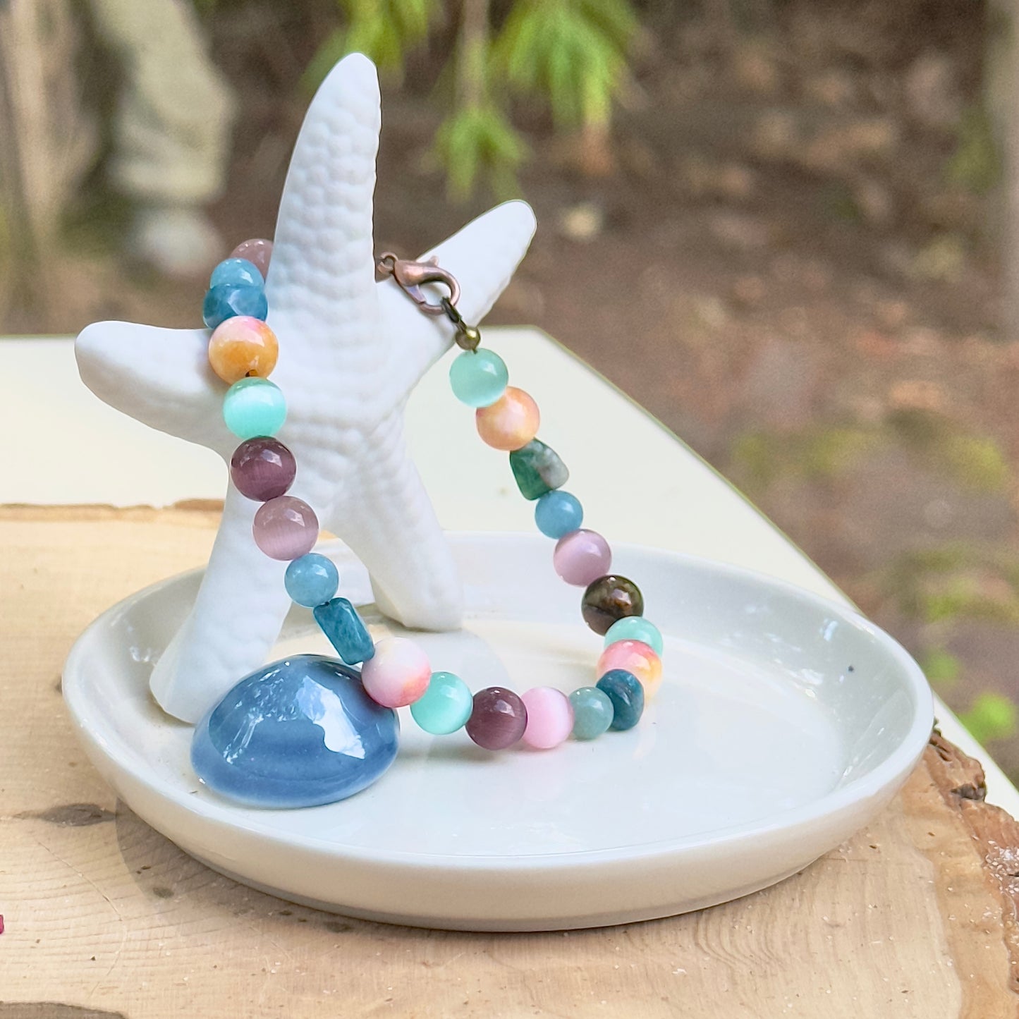 Handmade colorful gemstone bracelet displayed on a white starfish dish outdoors, styled with natural light and a calm, coastal setting.
