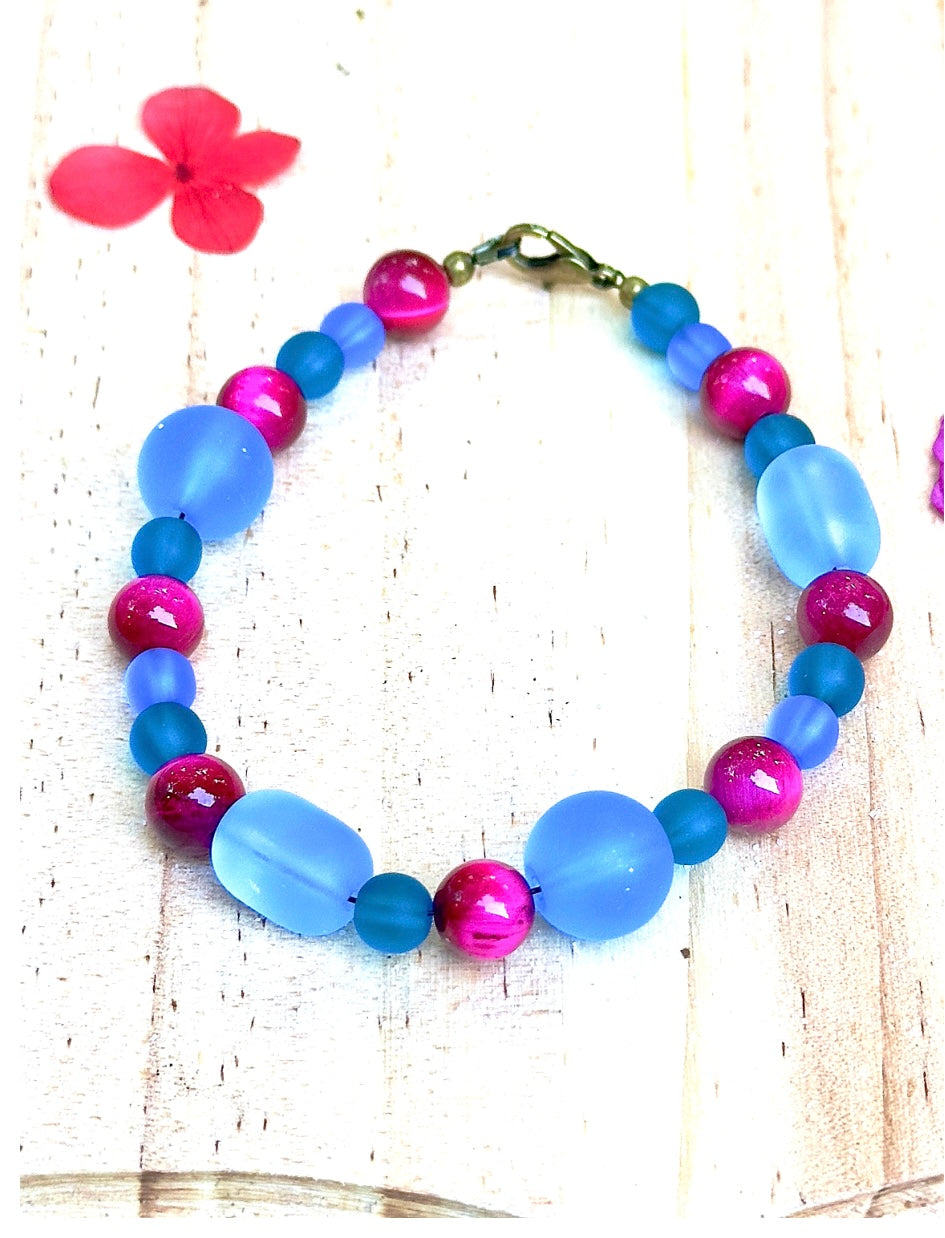Handmade beaded bracelet with frosted blue glass beads, teal accents, and pink marbled beads on a light wooden background.
