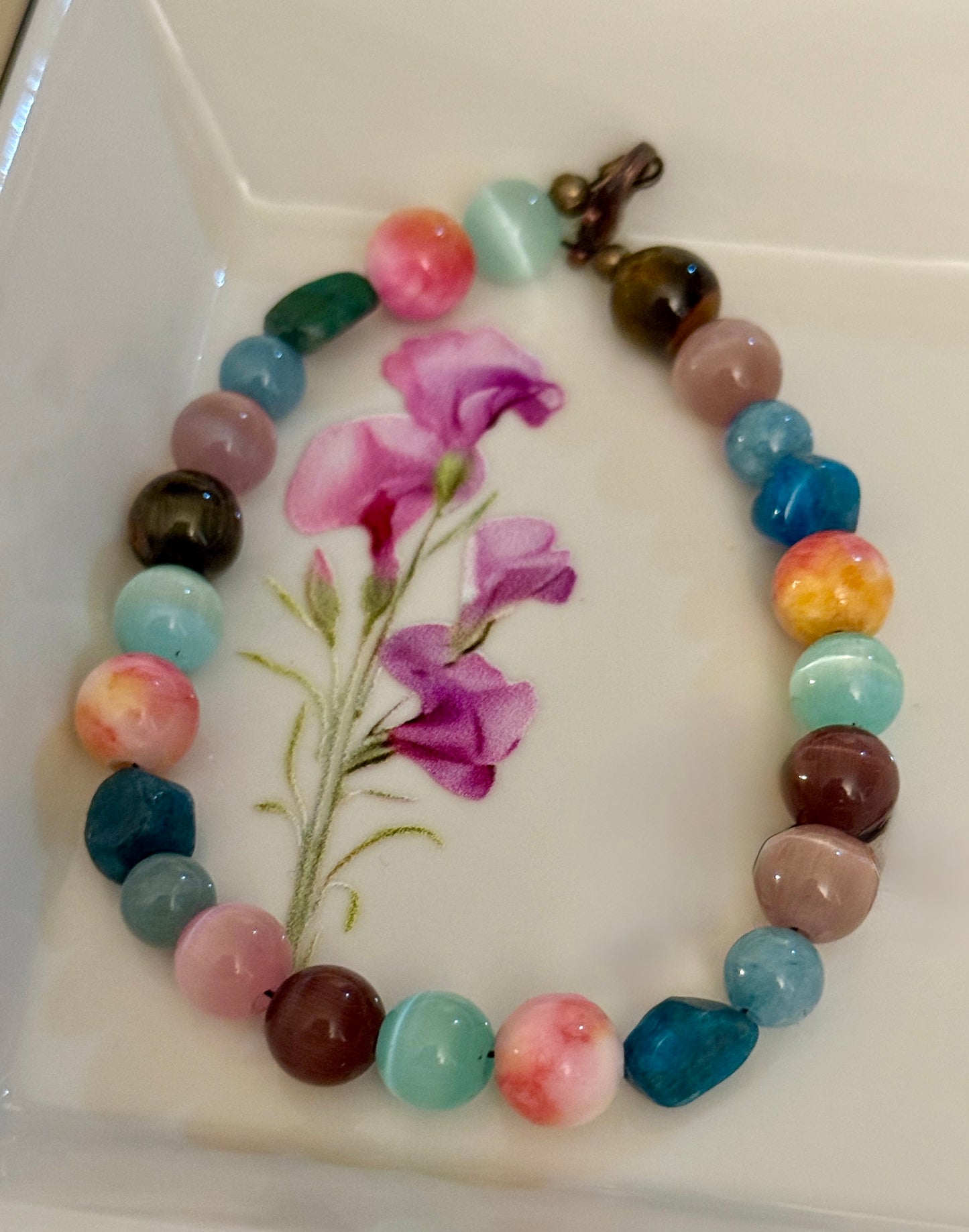 Colorful handmade gemstone bracelet arranged on a white dish with a pink floral design, showcasing pastel and earth-tone beads with a bronze clasp.