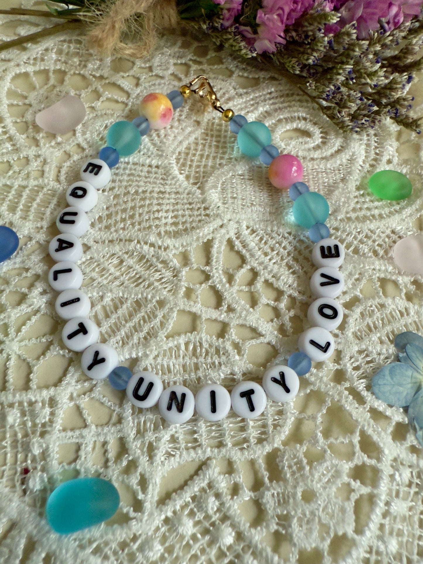 Handmade Equality Love Unity bracelet arranged on textured lace fabric with dried florals, highlighting soft ocean-toned beads.