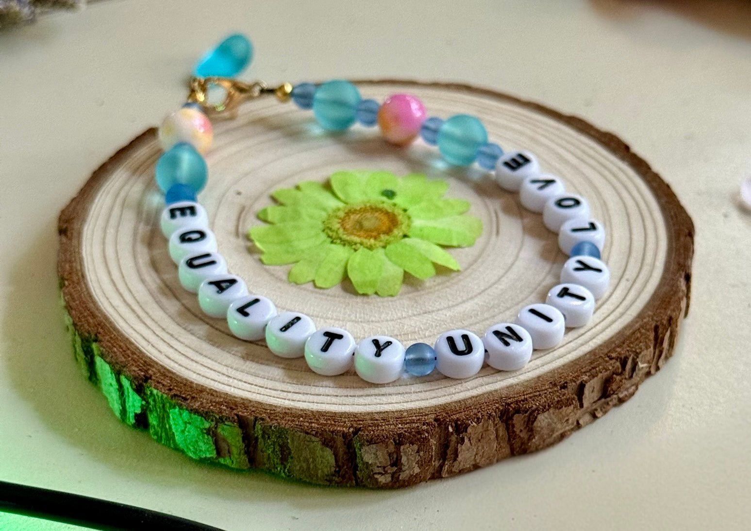 Equality Love Unity beaded bracelet on a wooden slice with green floral detail, styled in a gentle, earthy flat lay.