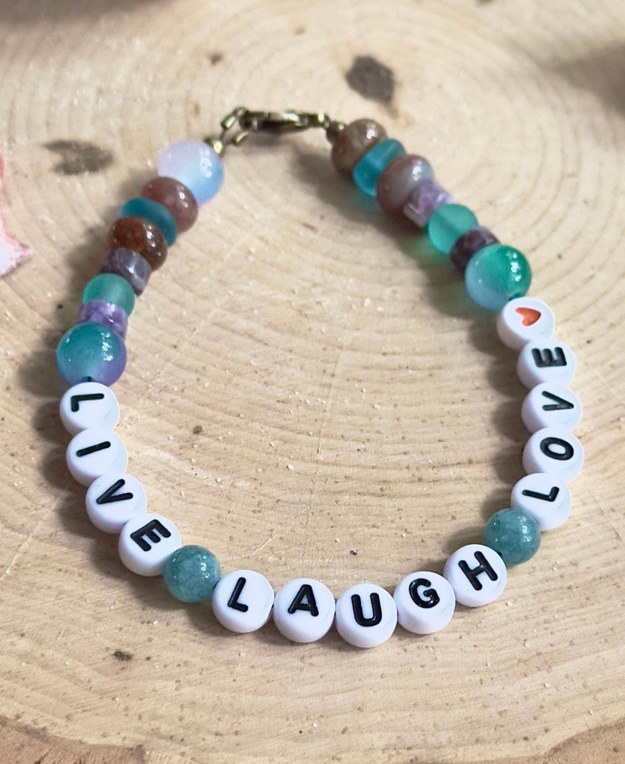 Handmade Live Laugh Love beaded bracelet displayed on a natural wood surface with turquoise, teal, and earth-toned gemstone beads.