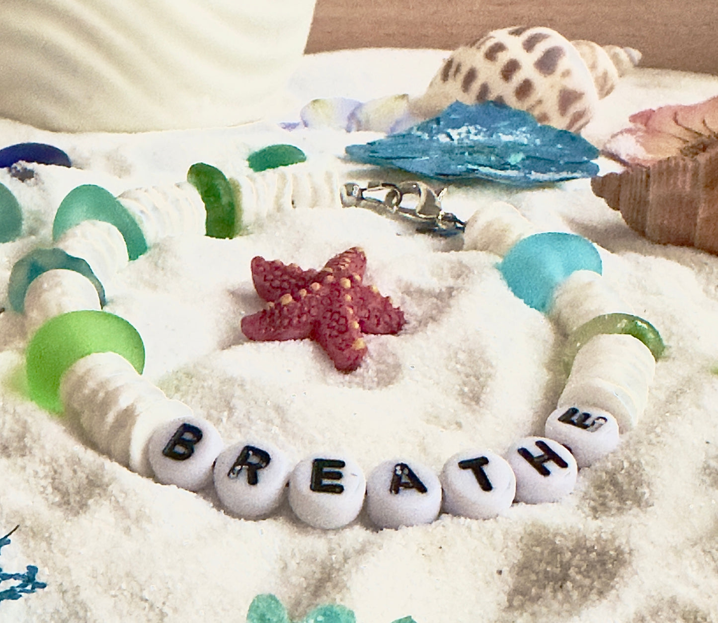 Ocean inspired Breathe bracelet with white beads, aqua and green accents styled in sand with coastal elements
