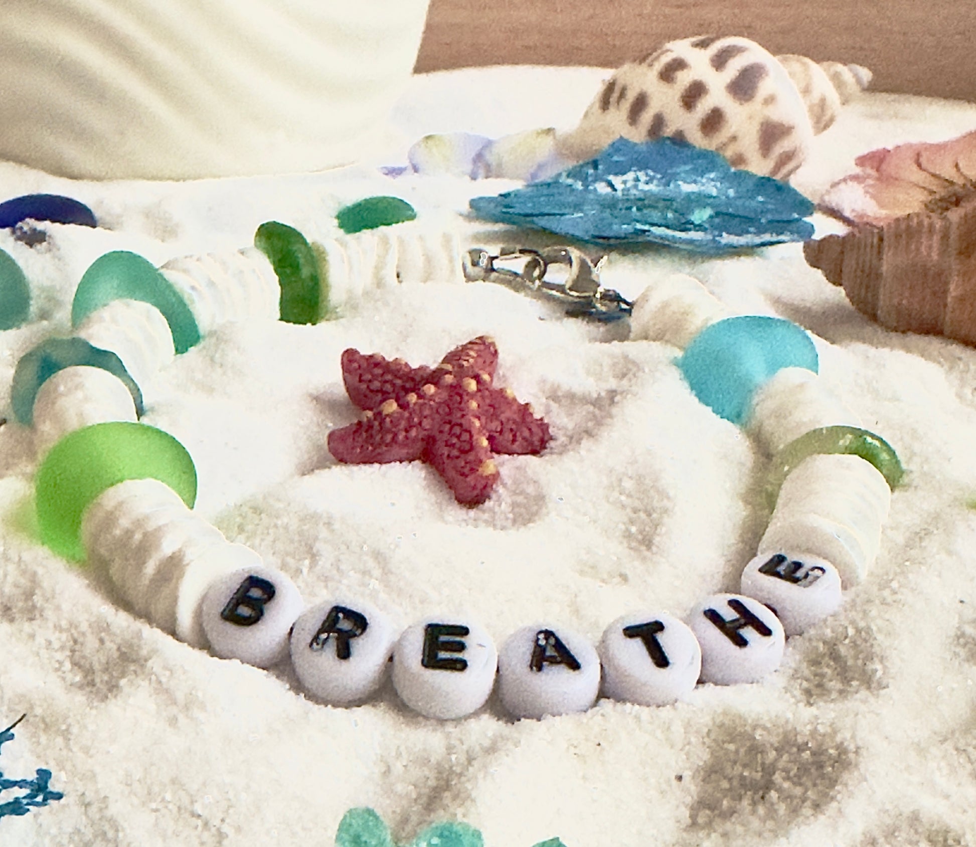 Ocean inspired Breathe bracelet with white beads, aqua and green accents styled in sand with coastal elements
