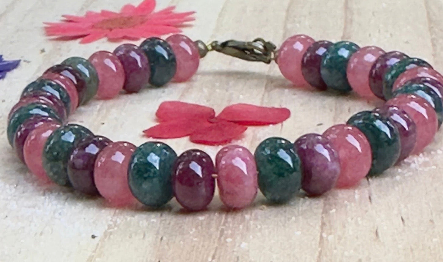 Between The Waves bracelet displayed on a white wooden surface with pink flower accents, showcasing colorful gemstone rondelle beads and bronze clasp.