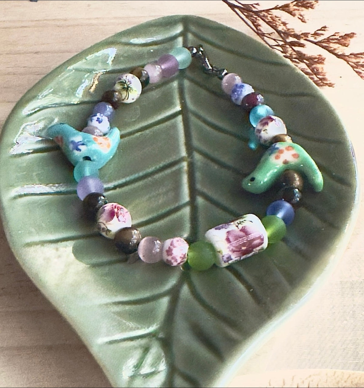 Bird-Eye View bracelet displayed on a green leaf-shaped dish, featuring floral beads and turquoise ceramic birds.