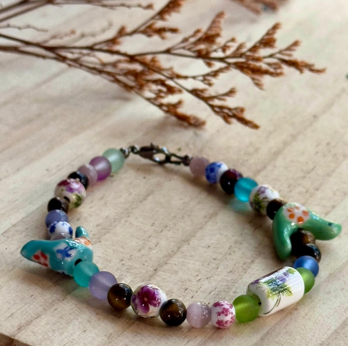 Bird-Eye View bracelet arranged on a light wood surface, showing colorful beads, floral details and turquoise bird accents.