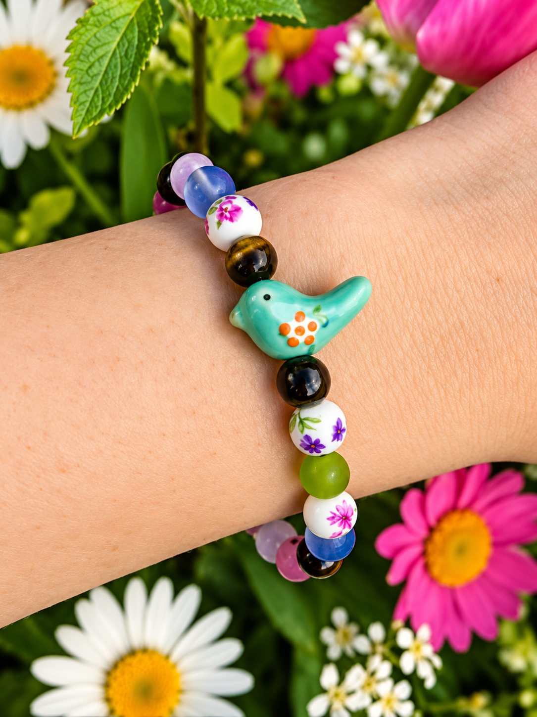     Bird-Eye View colorful bird and floral bracelet worn on a wrist against bright pink and white garden flowers.