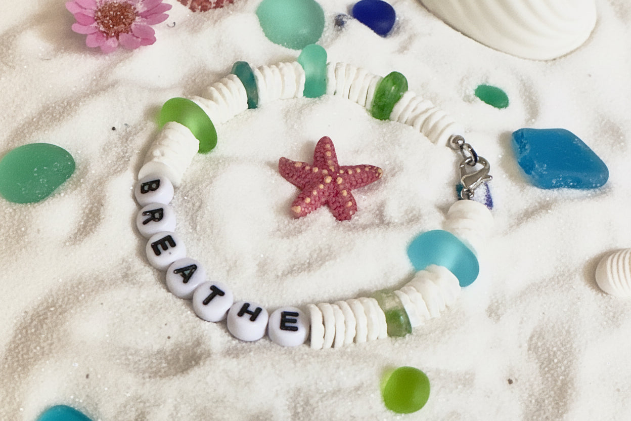 Bracelet with 'BREATHE' beads on a sandy surface with seashells and flowers.