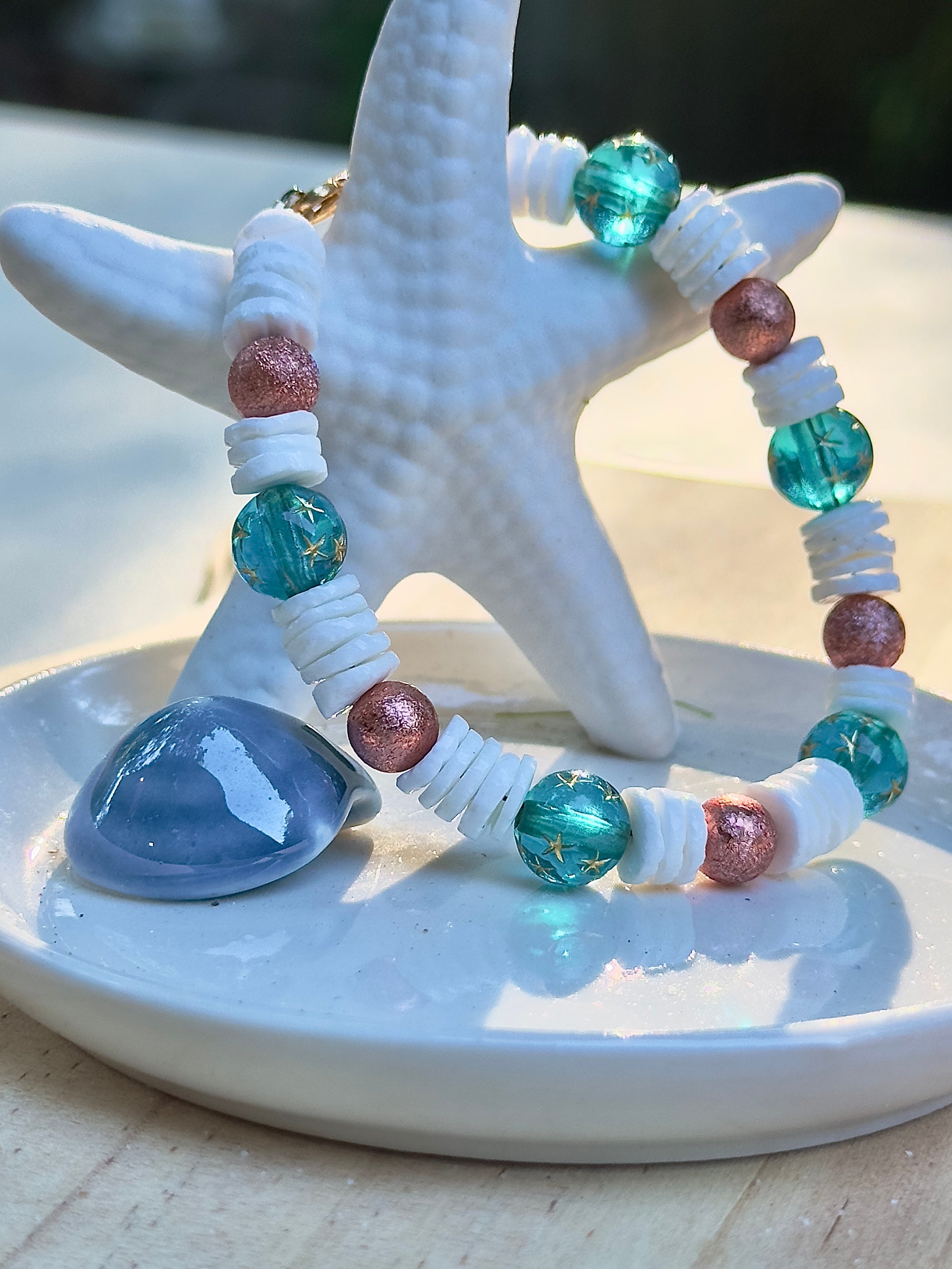 Handmade beaded bracelet with teal star beads and white shell spacers displayed on a ceramic starfish dish.
