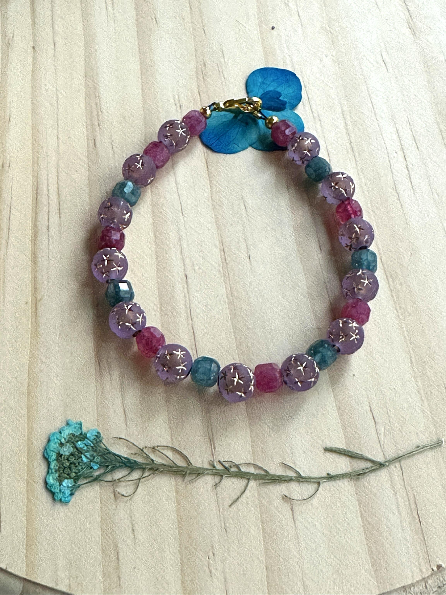 Handmade lavender and teal star gemstone bracelet displayed on a light wood background with a blue floral accent.