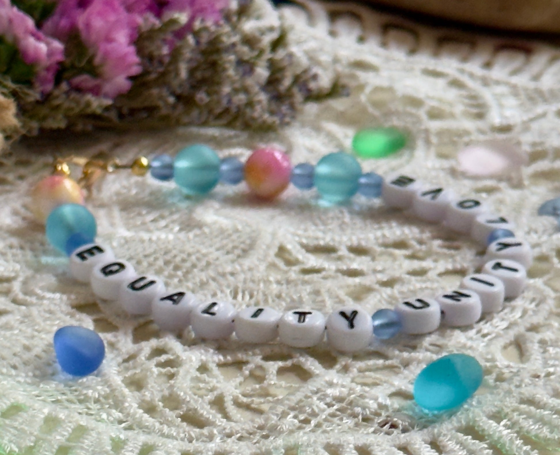 Close-up of Equality Love Unity bracelet beads showing pastel blue, aqua, and iridescent accents on lace fabric.