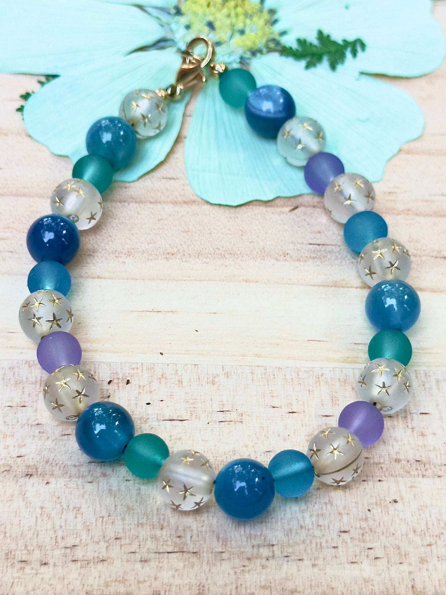 Beaded bracelet with blue, purple, and clear beads on a wooden surface with a shell and flower in the background.