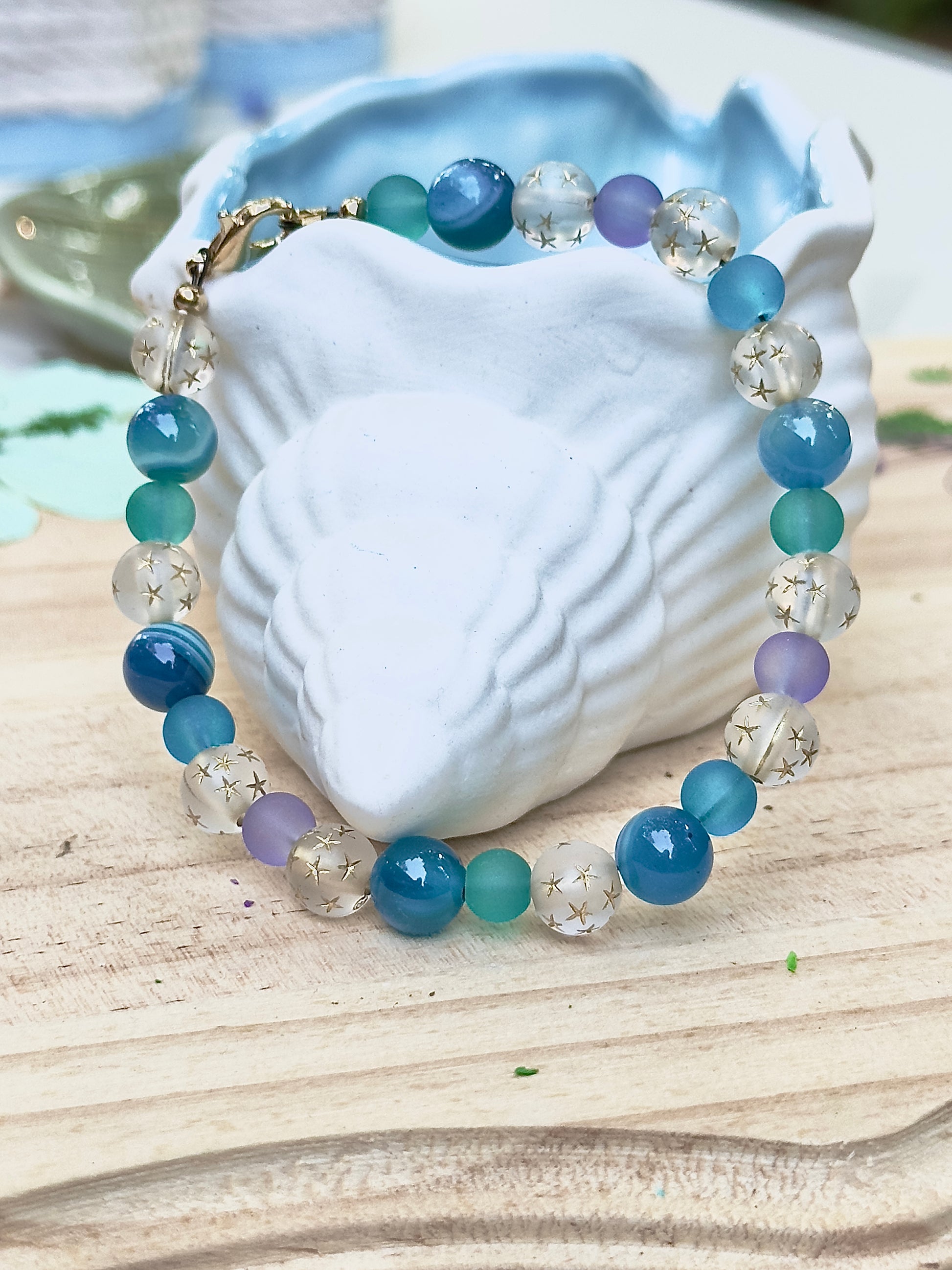 Celestial Opalite Sky bracelet styled on a white shell with blue and teal beads in a serene coastal-inspired setting