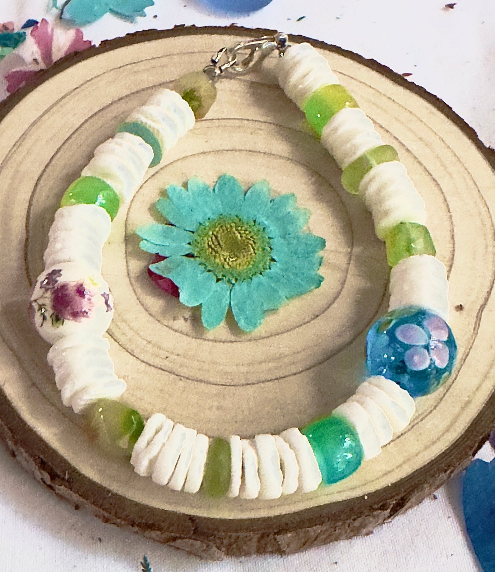 Nature-inspired bracelet with floral ceramic beads and green glass accents displayed on a wood slice.