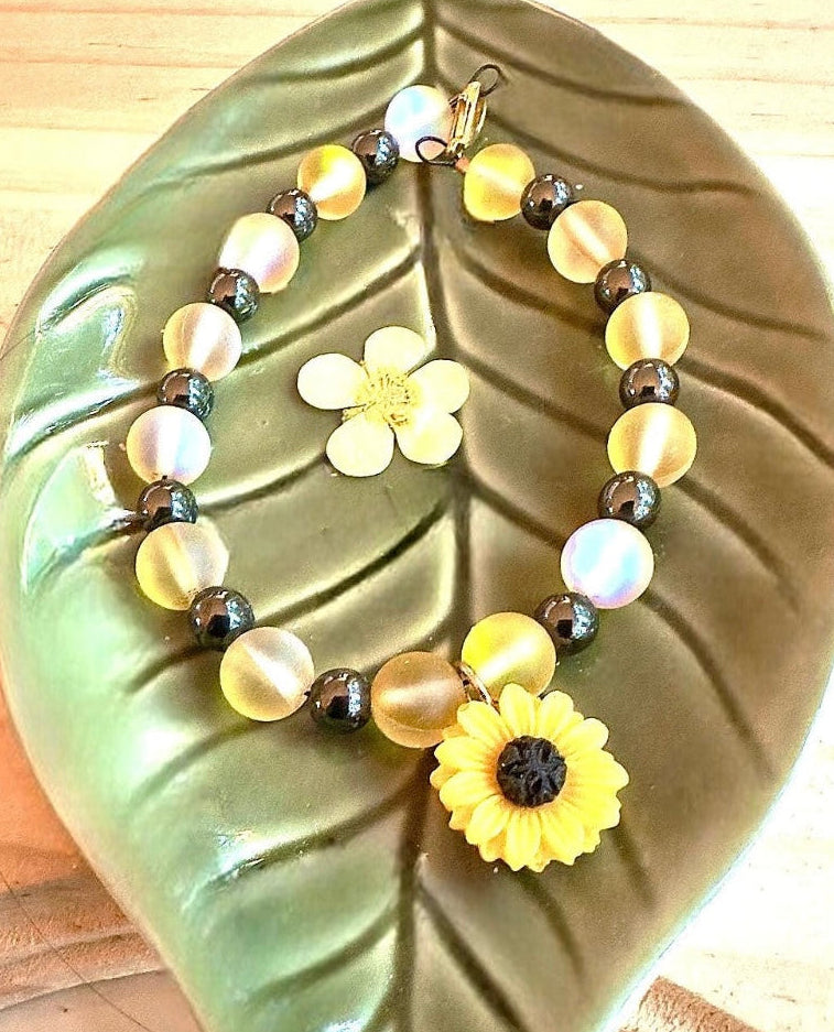 Close-up of a sunflower charm bracelet resting on a green leaf, highlighting frosted golden beads, metallic accents, and nature-inspired design details.
