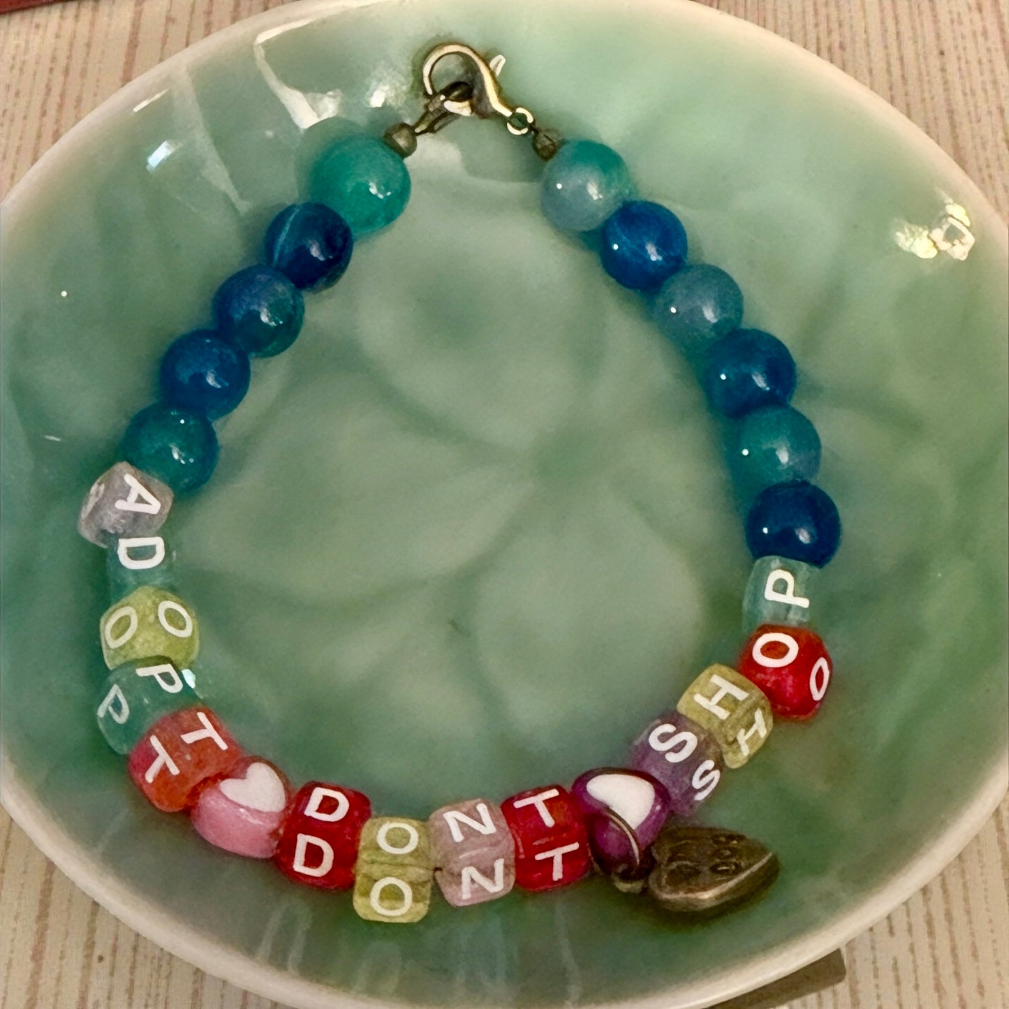 Adopt Don’t Shop beaded bracelet displayed in a soft teal ceramic bowl, highlighting gemstone colors, letter beads, and paw heart charm.
