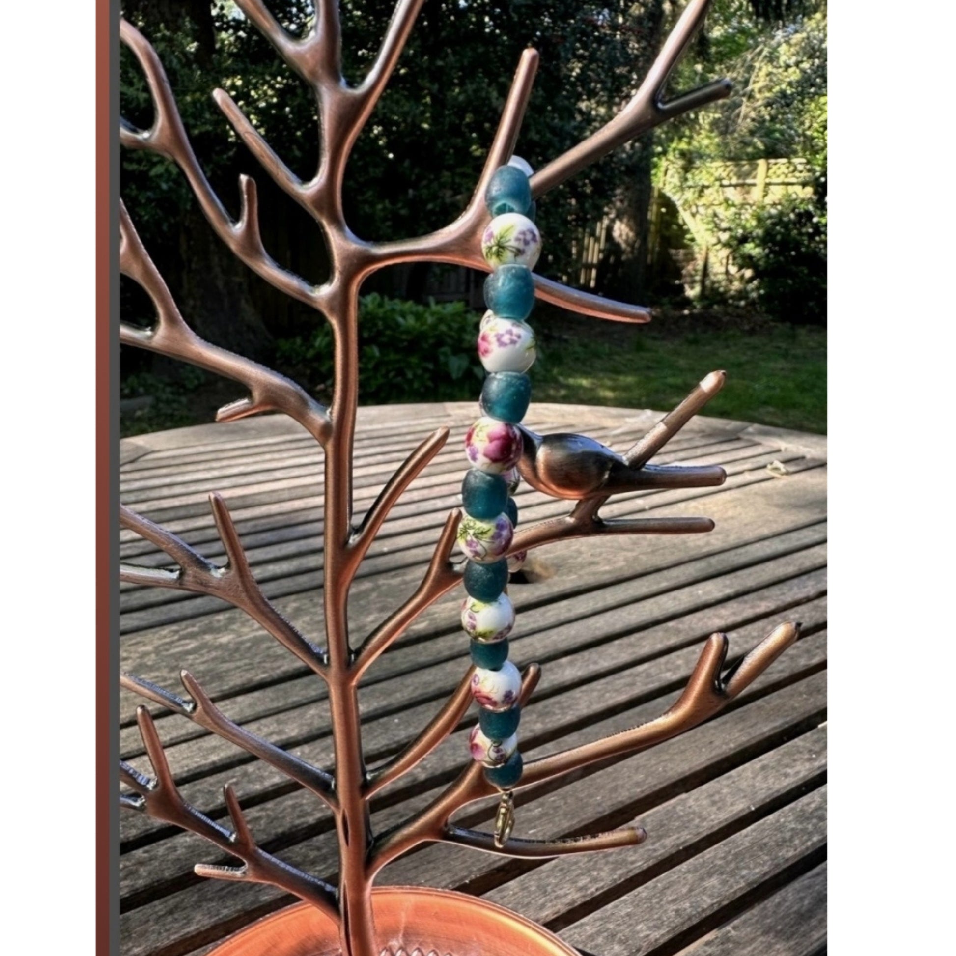 Nature-inspired handmade bracelet with floral ceramic and teal glass beads displayed on a copper jewelry tree, photographed outdoors in sunlight