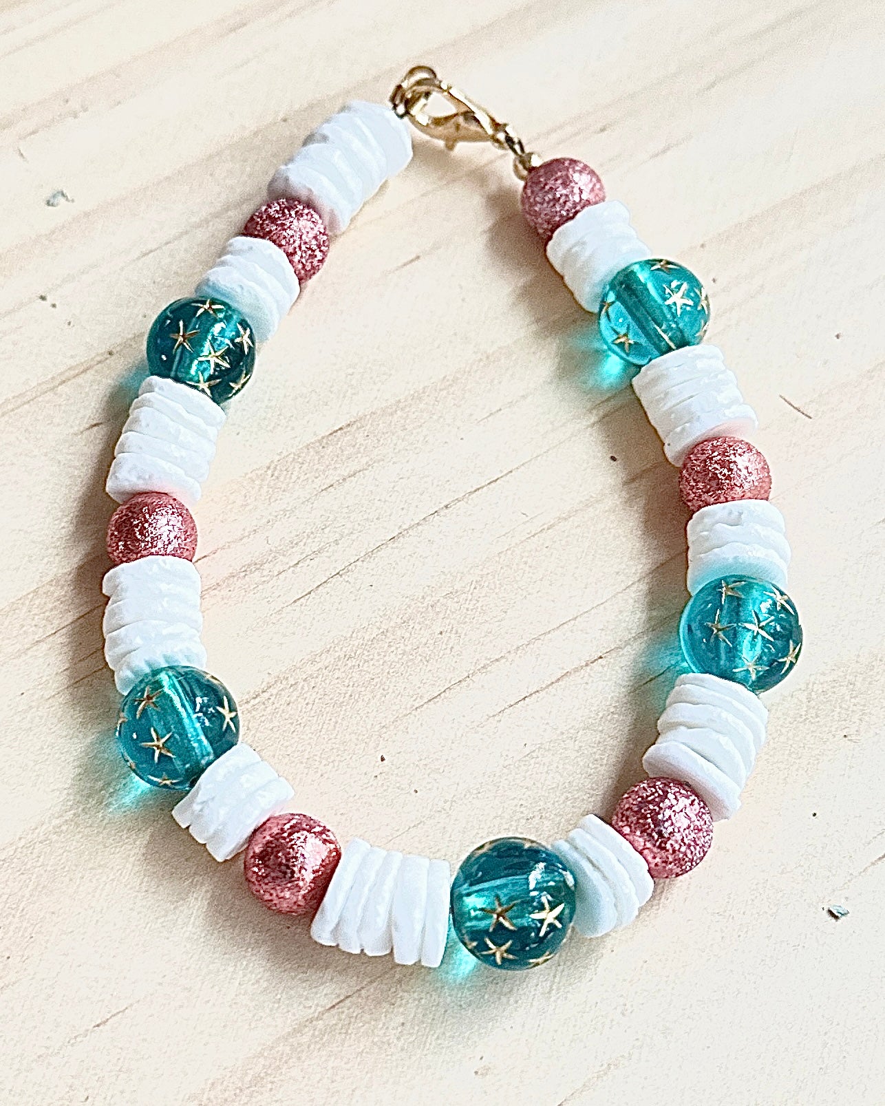 Handmade ocean-inspired beaded bracelet featuring teal star-pattern beads, white shell spacers, and rose shimmer accents on a neutral background.