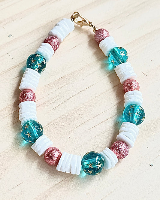 Handmade ocean-inspired beaded bracelet featuring teal star-pattern beads, white shell spacers, and rose shimmer accents on a neutral background.