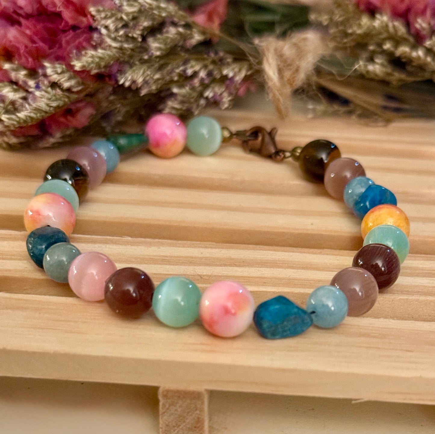 Handmade gemstone bracelet displayed on a natural wooden block, featuring soft pastel pinks, blues, greens, and grounding earth tones.