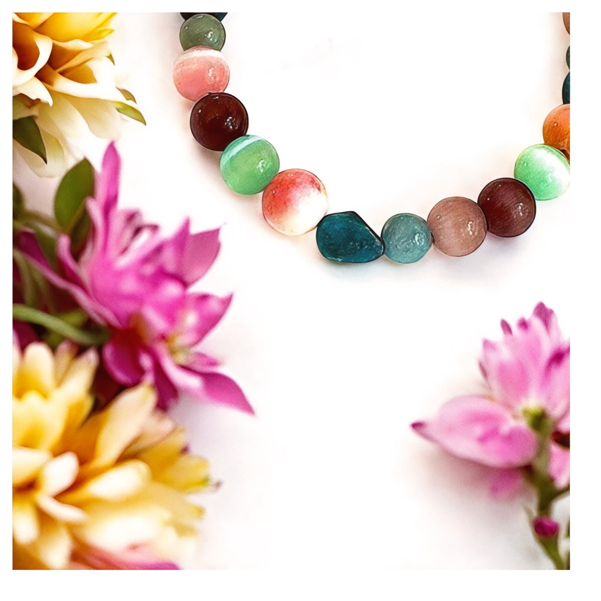 Close-up of a colorful gemstone bracelet resting among pink and yellow flowers on a white background, highlighting the texture and pastel tones of the beads.