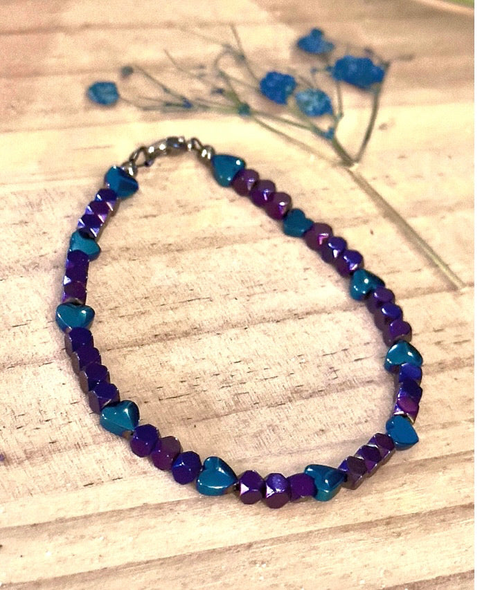 Heartspell Twilight purple and blue beaded bracelet with turquoise heart accents displayed on a light wood surface.